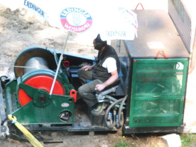 Dieses Bild zeigt die Antriebsstation der Bauseilbahn im Bereich der Bergstation der Grenzwiesbahn. Mit der Seilrolle links wird das Zugseil eingeholt. Der Arbeiter, der die Bahn bedient, sitzt im Freien unter einem Sonnenschirm. Insgesamt macht das Ganze einen sehr improvisierten Eindruck. Aber es funktioniert!