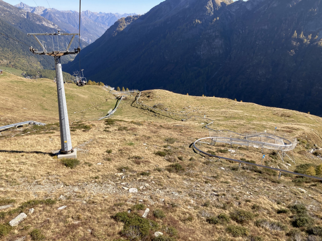 Blick auf die kurze Rodelbahn. Wir machten eine Fahrt für 10 Franken. Fazit: Nicht lohnenswert. Erstens ist die Bahn sehr kurz. Ok. Zweitens gibt es bei bei jeder Kurve üble Schläge in den Rücken und an ein dosiertes Abbremsen bzw. Beschleunigen ist nicht zu denken. Bei jeder kleinsten Bremseinleitung erfolgt quasi eine Vollbremsung. <br />Die Rodelbahn benötigt viel Personal: Eine Kassiererin, die auch die Instruktion erteilt. Dann eine Person am unteren Bahnende, welche den Schlitten auf die Bergförderanlage schiebt (die automatische Vorrichtung hat nicht funktioniert). Und weiter ein Angestellter nach der Bergfahrt, der den Bob wieder zum Start schiebt (auch hier hat die Technik nicht funktioniert). So kann das nicht rentabel sein...