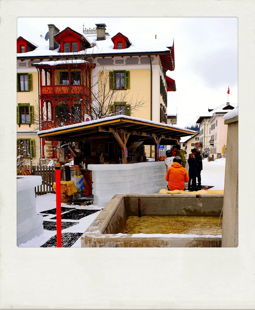 Biegt man nach dem letzten Viadukt unter der Albulabahn nach rechts ins Dorf ab und nicht nach links zum Sessellift, fährt man zwischen Ställen und vorbei an der Dorfkirche die Hauptstraße hinab zu dieser Eisbar. Hier kann man ebenfalls ganz entspannt einen Après-Ski-Drink nehmen und wieder mit den Barkeepern plaudern, die u.a. vom Bau der Eisbar berichten: Man ließ Wasser ins Bergüner Freibad laufen, einfrieren und schnitt dann Blöcke heraus und stapelte sie hier am Dorfbrunnen auf. Ein Block wiegt um die 80kg. Schöne Idee und mit Herzblut gemacht! Stolz waren sie insbesondere auf die nächtliche Beleuchtung.